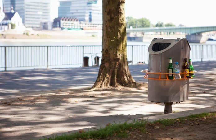 Ein orangener Pfandring ist um einen Mülleimer an einem Fußweg "geschnallt".