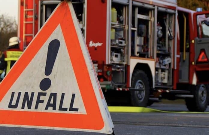 Dreieckiges Warnschild mit der Aufschrift „Unfall!“ im Vordergrund, im Hintergrund ein Einsatzfahrzeug der Feuerwehr mit geöffneten Fächern und Ausrüstung.
