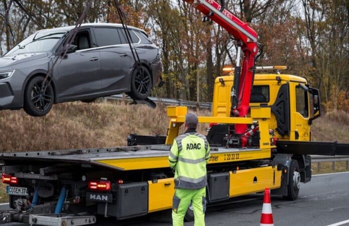 Ein ADAC-Abschleppdienst hebt mit einem Kran einen grauen Kombi auf einen gelben Abschleppwagen nach einem Unfall oder einer Panne auf der Autobahn. Ein ADAC-Mitarbeiter in Warnkleidung steht daneben und überwacht den Vorgang. Im Hintergrund herbstliche Bäume und eine Leitplanke. Sicherer Fahrzeugtransport und Pannenhilfe auf deutschen Straßen.