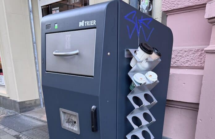 Smarte Mülltonne der Stadt Trier mit LED-Anzeige und seitlicher Halterung, gefüllt mit benutzten Kaffeebechern. Die Tonne zeigt sichtbare Gebrauchsspuren und Graffiti an der Seite