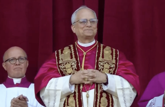 Papst Leo der 14. nach seiner Wahl auf dem Balkon des Petersdoms in Rom, mit segnender Geste vor rotem Vorhang, flankiert von Geistlichen.