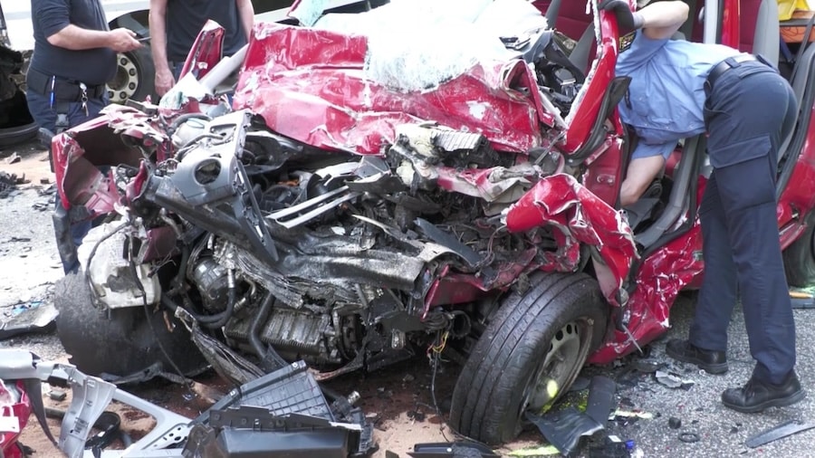 Verformte Fahrzeugfront eines roten Vans nach Frontalunfall mit LKW, Polizisten arbeiten an der Unfallstelle, Trümmer auf der Straße.