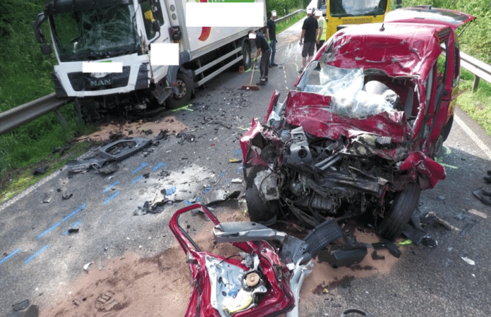 Frontansicht eines roten Fahrzeugs mit komplett zerstörtem Motorblock nach Kollision mit weißem LKW, Trümmer auf der Straße verteilt.