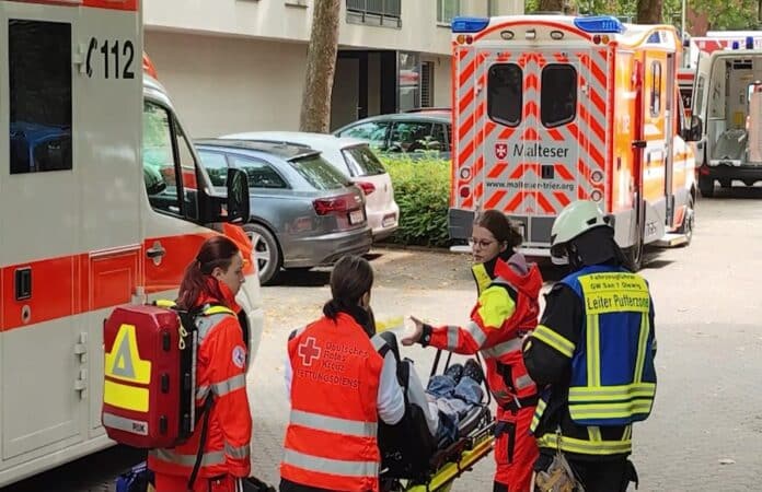Einsatzkräfte des Deutschen Roten Kreuzes und der Feuerwehr versorgen eine Person auf einer Trage vor einem Rettungswagen in einem Wohngebiet nahe der Schule.