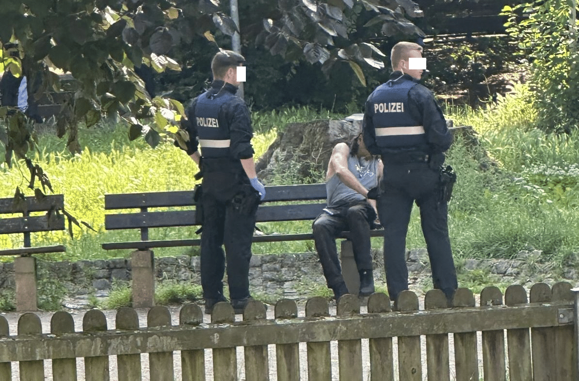 Zwei Polizisten kontrollieren einen auf einer Parkbank sitzenden Mann.
