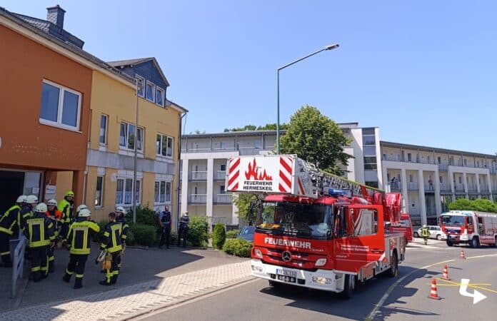 Feuerwehrleute in Schutzkleidung vor einem Wohnhaus in Hermeskeil, Drehleiter und Einsatzfahrzeuge im Hintergrund, Notfalleinsatz in städtischer Umgebung.
