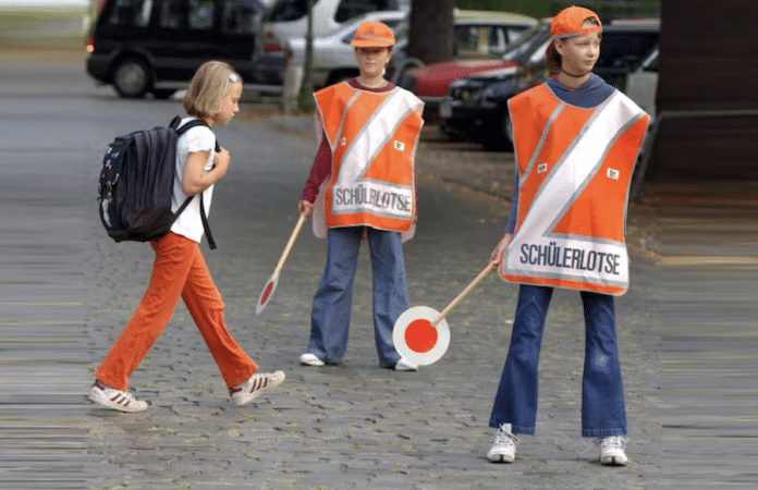 Zwei Schülerlotsen in orangefarbener Warnkleidung helfen einem Schulkind beim sicheren Überqueren der Straße. Foto: dpa